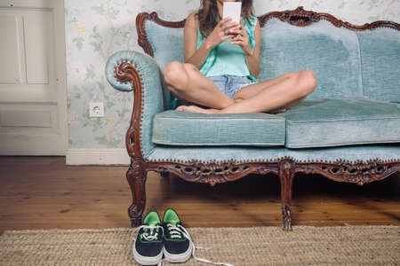 Closeup of beautiful young woman looking smartphone sitting on a classic sofa with legs crossed and her sneakers over the carpetの写真素材