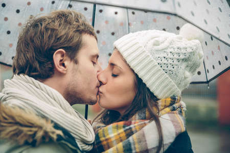 Closeup of young beautiful couple kissing under the umbrella in an autumn rainy day. Image focused on the lips.の写真素材