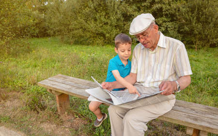 Senior man reading newspaper and cute child pointing an article with his finger sitting on park...