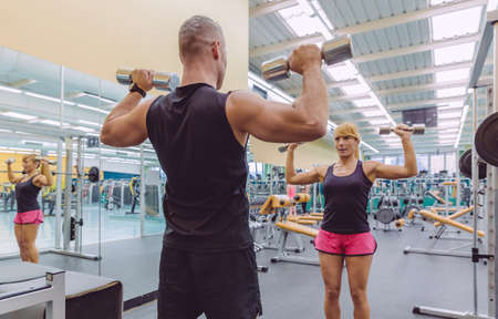Man coach training to beautiful woman with dumbbells exercises on fitness center. Personal trainer...