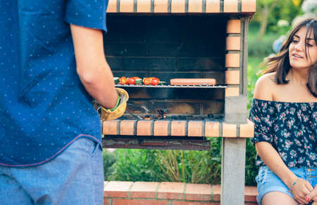 Back view of young man cooking sausages and vegetable skewers in a brick barbecue while woman looking in a outdoors summer partyの写真素材