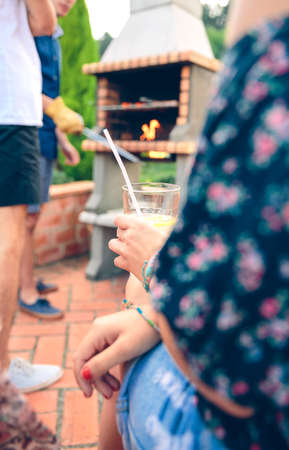 Closeup of young woman sitting and holding lemonade glass in her hands with friends in the background cooking and talking in a outdoors summer barbecueの写真素材