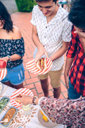 Happy young man holding an american hot dog in a outdoors summer barbecue with his friendsの写真素材