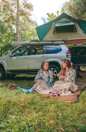 Happy young women friends resting sitting under a blanket and their 4x4 vehicle with tent in the roof on the backgroundの写真素材