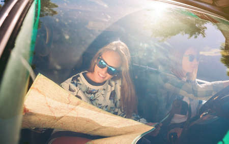 Portrait of happy young woman with sunglasses looking a map inside of car while her friend driving in a road trip adventure. Female friendship and leisure time concept.の写真素材