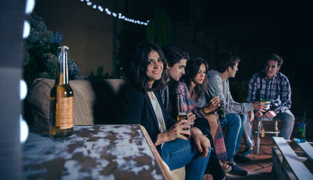 Portrait of smiling young woman sitting and holding a beer while her friends talking in a outdoors party. Friendship and celebrations concept.の写真素材