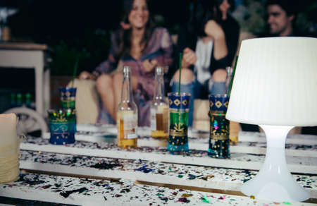 Closeup of white lamp over a pallets table with drinks and confetti in a outdoors party with people talking in the background. Selective focus on lamp.の写真素材