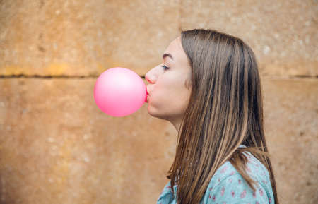 Closeup of beautiful young brunette teenage girl blowing pink bubble gum over a stone wall backgroundの写真素材