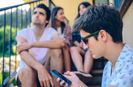 Portrait of young man looking a smartphone outdoors sitting on the home stairs steps with his friends. Young people lifestyle concept.の写真素材