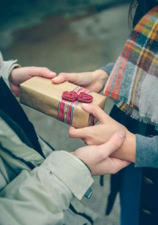 Closeup of man hands giving a gift box with red handmade flowers to woman with scarf in a cold autumn day. Love and couple relationships concept.の写真素材