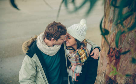 Portrait of young couple in love with hat and scarf embracing and kissing under a tree in a cold...