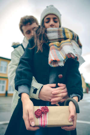 Protrait of young beautiful woman showing gift box in her hands and her boyfriend embracing from behind outdoors in a cold autumn day. Love and couple relationships concept.の写真素材