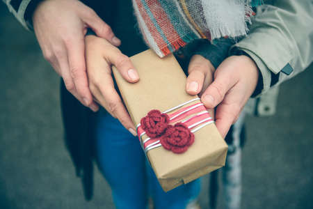 Closeup of woman and man hands showing a gift box with red handmade flowers otdoors in a cold autumn day. Love and couple relationships concept.の写真素材