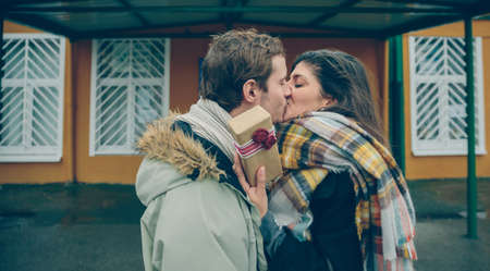 Young beautiful woman with gift box in the hand embracing and kissing to her boyfriend for the surprise outdoors in a cold autumn day. Love and couple relationships concept.の写真素材