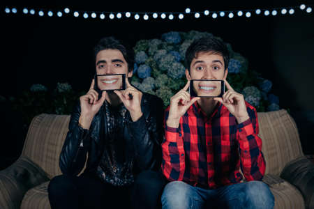 Closeup of two funny young men holding smartphones over their faces showing female mouths smiling in the screen on a outdoors party. Friendship and celebrations concept.の写真素材