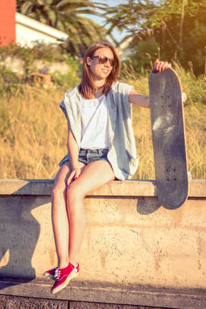 Portrait of beautiful young girl with short shorts and skateboard sitting over the wall on a summer day. Warm tones edition.の写真素材
