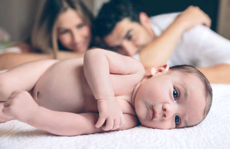 Portrait of peaceful newborn lying over a bed and young couple looking and smiling on the background. Family and baby care concept.の写真素材