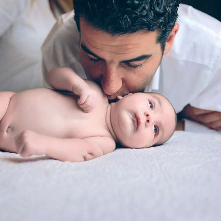 Young handsome man kissing in the neck to peaceful newborn lying over a bed. Paternity and baby care...