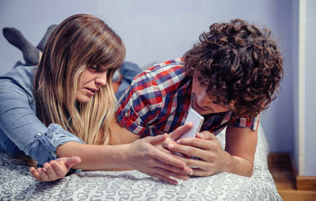 Young man looking private messages in a smartphone of angry woman lying over a bed. Bullying and...