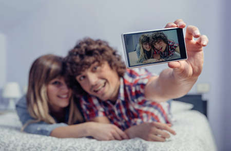 Happy young couple in love taking a selfie with smartphone lying over a bed. Selective focus on the...