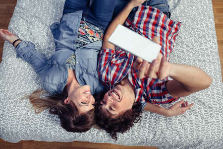 Top view of happy young couple in love taking a selfie with smartphone lying over a bed. Leisure time at home concept.の写真素材