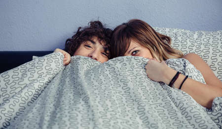 Happy young couple in love laughing and covering their mouths under a duvet. Love and couple relationships concept.の写真素材