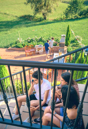 Young man calling by phone sitting on the home stairs with his friends over a summer barbecue background. Young people lifestyle concept.の写真素材