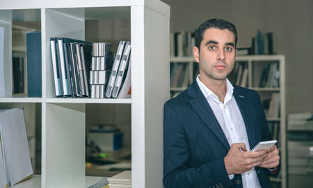 Portrait of handsome businessman holding smartphone leaning on a bookcase