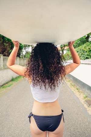 Back view of curly brunette girl with white top and black bikini holding surfboard over head on the street. Summer lifestyle concept.の写真素材