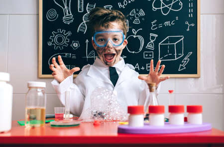 Portrait of happy little scientist with glasses and dirty face opening his arms behind of glass with soap foam over tableの写真素材