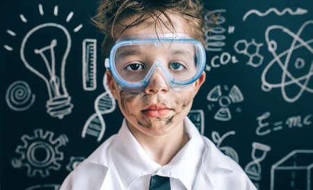 Close up of serious scientist boy with glasses and dirty face looking at camera against of chalkboard with drawingsの写真素材