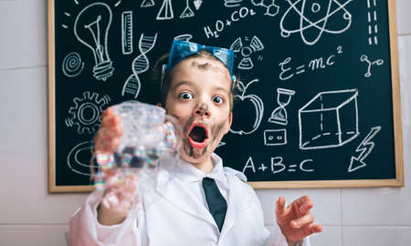 Close up of excited little scientist with dirty face holding glass with soap foam against of blackboard with drawingsの写真素材