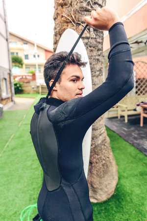 Portrait of young surfer man with surfboard closing his wetsuit in front of palm tree. Summer sports concept.の写真素材