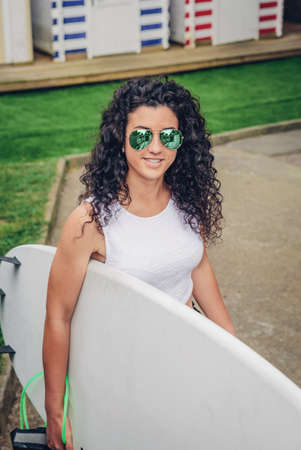 Closeup of of curly brunette surfer woman with white top and sunglasses walking with a surfboardの写真素材