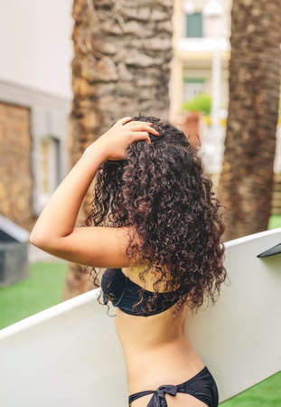 Back view of curly brunette surfer woman with black bikini holding a surfboard over a palm tree backgroundの写真素材