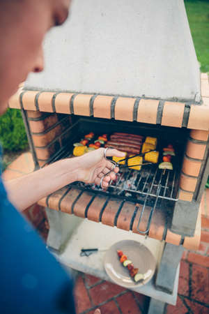 Back view of unrecognizable young man cooking sausages, corn and vegetable skewers in a brick barbecueの写真素材