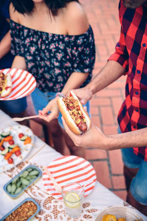 Close up of unrecognizable young man holding an american hot dog in a outdoors summer barbecue with his friendsの写真素材