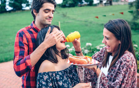 Funny girl pouring mustard pot inside of woman mouth while man closing their eyes with hands in a summer party.の写真素材