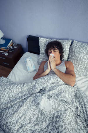 Portrait of young man sneezing and covering nose with tissue lying on bed. Sickness and healthcare concept.の写真素材