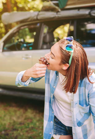 Portrait of beautiful young woman with plaid shirt eating chocolate ice cream against of car in forestの写真素材