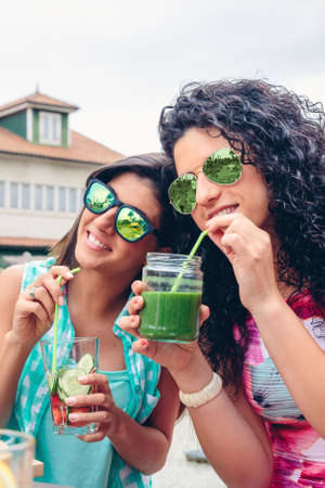 Two young women with sunglasses drinking healthy organic beverages with straws outdoorsの写真素材