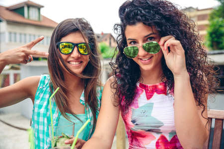 Close up of two happy young women with sunglasses smiling and looking at camera while holding green vegetable smoothies outdoorsの写真素材
