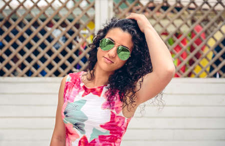 Portrait of curly young woman with sunglasses looking at camera over white garden fence background