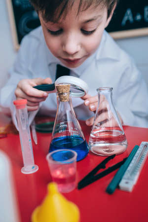 Close up of happy little scientist looking colorful chemical liquid in flask through magnifier against of drawn blackboardの写真素材