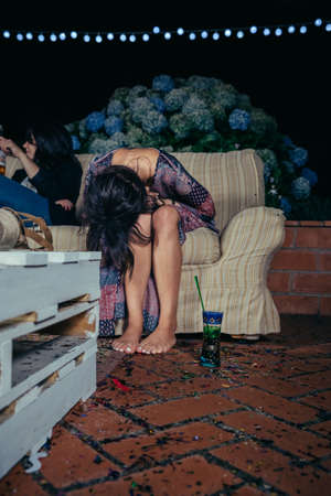Young drunk woman holding her head sitting in the sofa in a outdoors party. Fun and alcohol and drugs problems concept.の写真素材