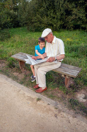 Senior man and cute child reading a newspaper sitting on park bench. Two different generations concept.の写真素材
