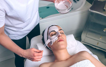 Female beautician applying clay facial mask to young woman lying with closed eyes in spa. Medicine,...