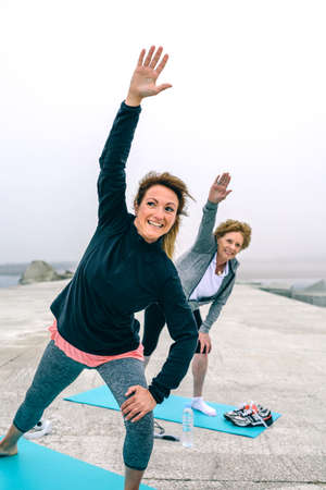 Happy personal trainer with senior woman stretching side by sea pierの写真素材