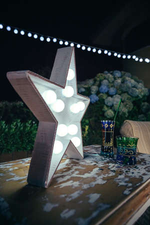 Close up of white handmade star lamp with light bulbs over a wooden table with drinks and lights garland in the background in a outdoors party.の写真素材