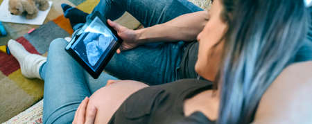Pregnant woman with her partner looking in the tablet a ultrasound of their baby in the living roomの写真素材